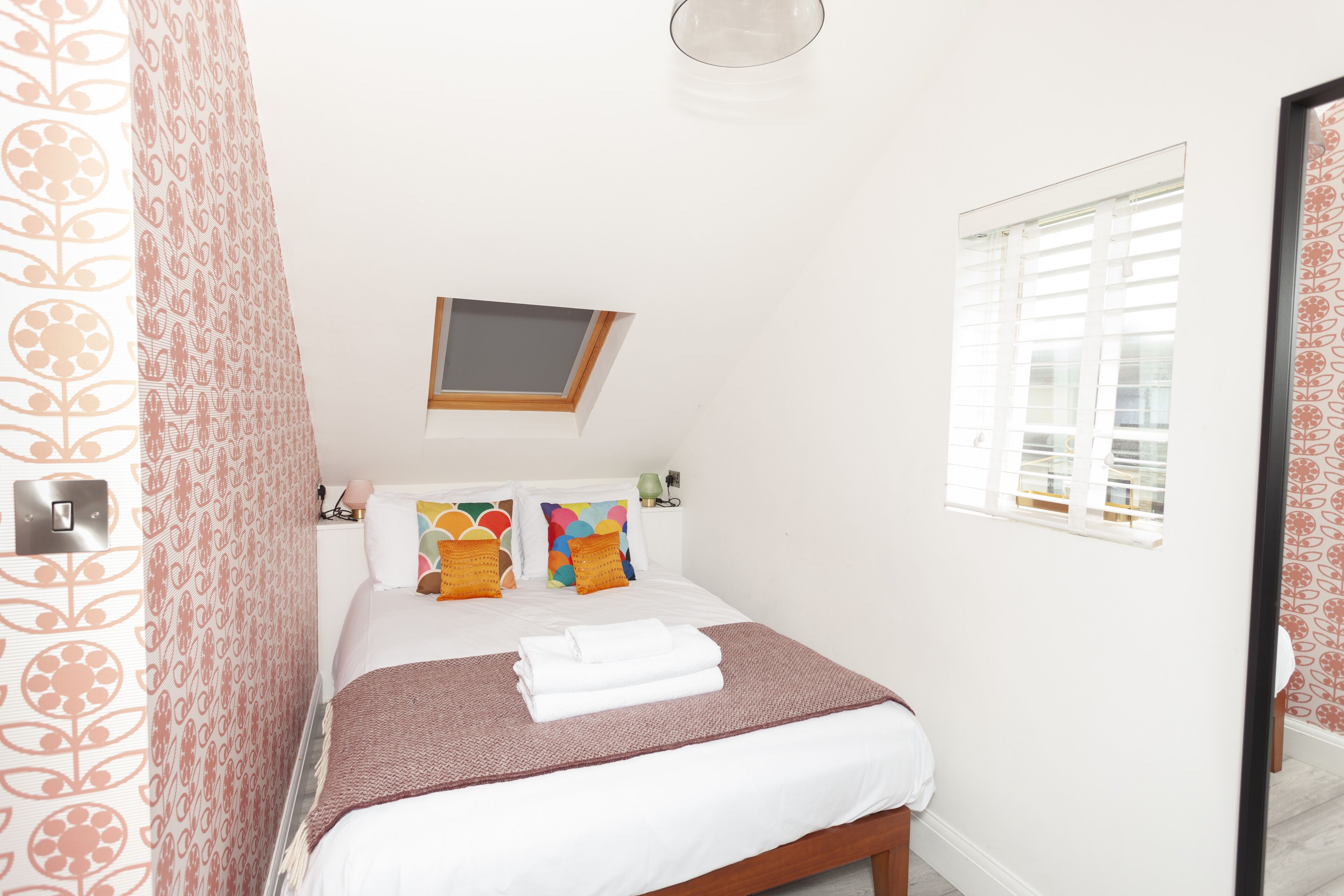 Een keurig opgemaakt bed met een bruine sprei en witte lakens, een raam met zonwering en een wandlamp.