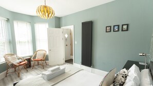 A bedroom with a bed, two wicker chairs, a radiator, and a door leading to another room. - Sunbeam Cottage (Whitstable)