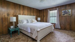 Quarto com parede revestida de madeira, cama branca com cabeceira, duas mesas de cabeceira com abajures, janela com cortinas e um quadro na parede.