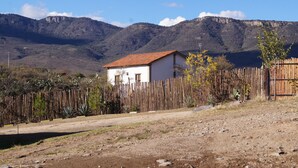 Uma casa rústica com chão de terra e cerca de madeira em uma região montanhosa.
