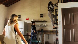 Uma cozinha com uma pessoa subindo em uma prateleira, uma sala de jantar com mesa e cadeiras, e uma porta à direita.