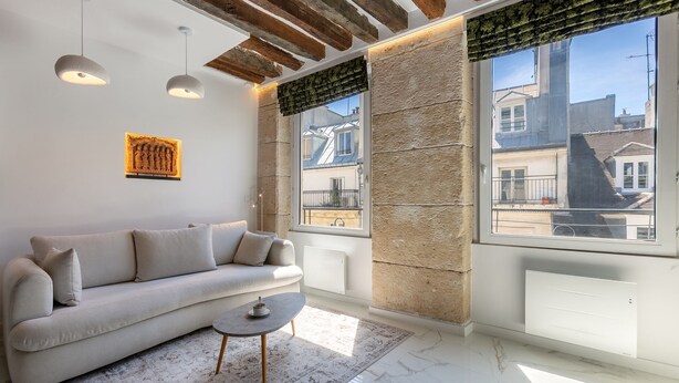 A modern living room with a sofa, a coffee table, and a view of buildings through large windows. - Parisian Pied-a-terre in the heart of the city - Close to everything (Paris)