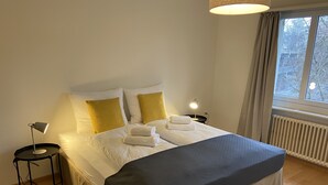 A neatly made bed with white and yellow pillows, a black bedside table with a lamp, and a window with curtains. - K190 - 2BDR Apartment close to Zurich Airport (Zürich)