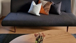 A modern living room with a dark grey sofa, a round wooden coffee table, and a wall-mounted abstract art piece. - K190 - 2BDR Apartment close to Zurich Airport (Zürich)