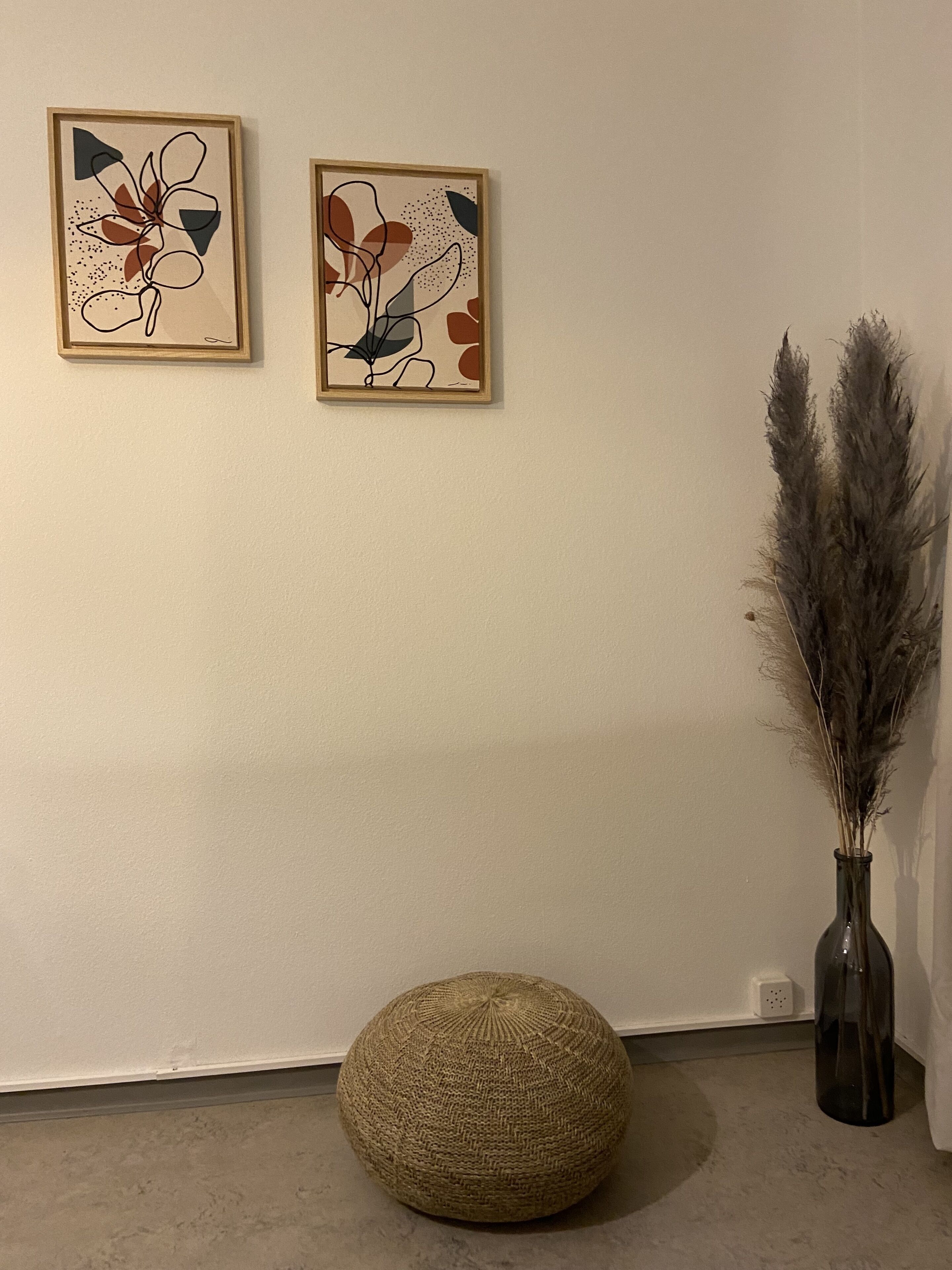 A room with two abstract art pieces on the wall, a tall vase with pampas grass, and a round woven ottoman on the floor.