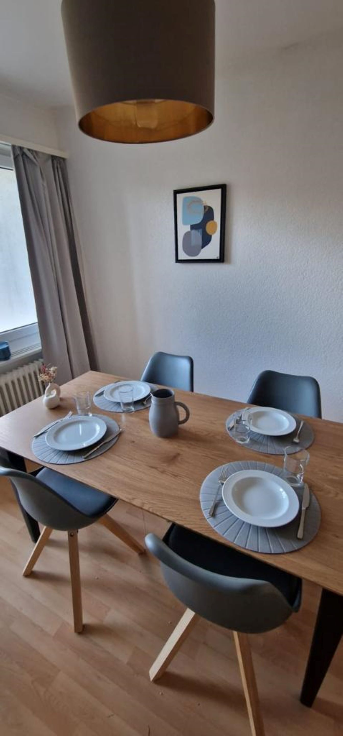 Zona pranzo con tavolo in legno apparecchiato per quattro persone, piatti e caraffa grigi. Due sedie, una con cuscino nero. Sopra il tavolo pende una lampada a sospensione e al muro è appesa un'opera d'arte astratta incorniciata.