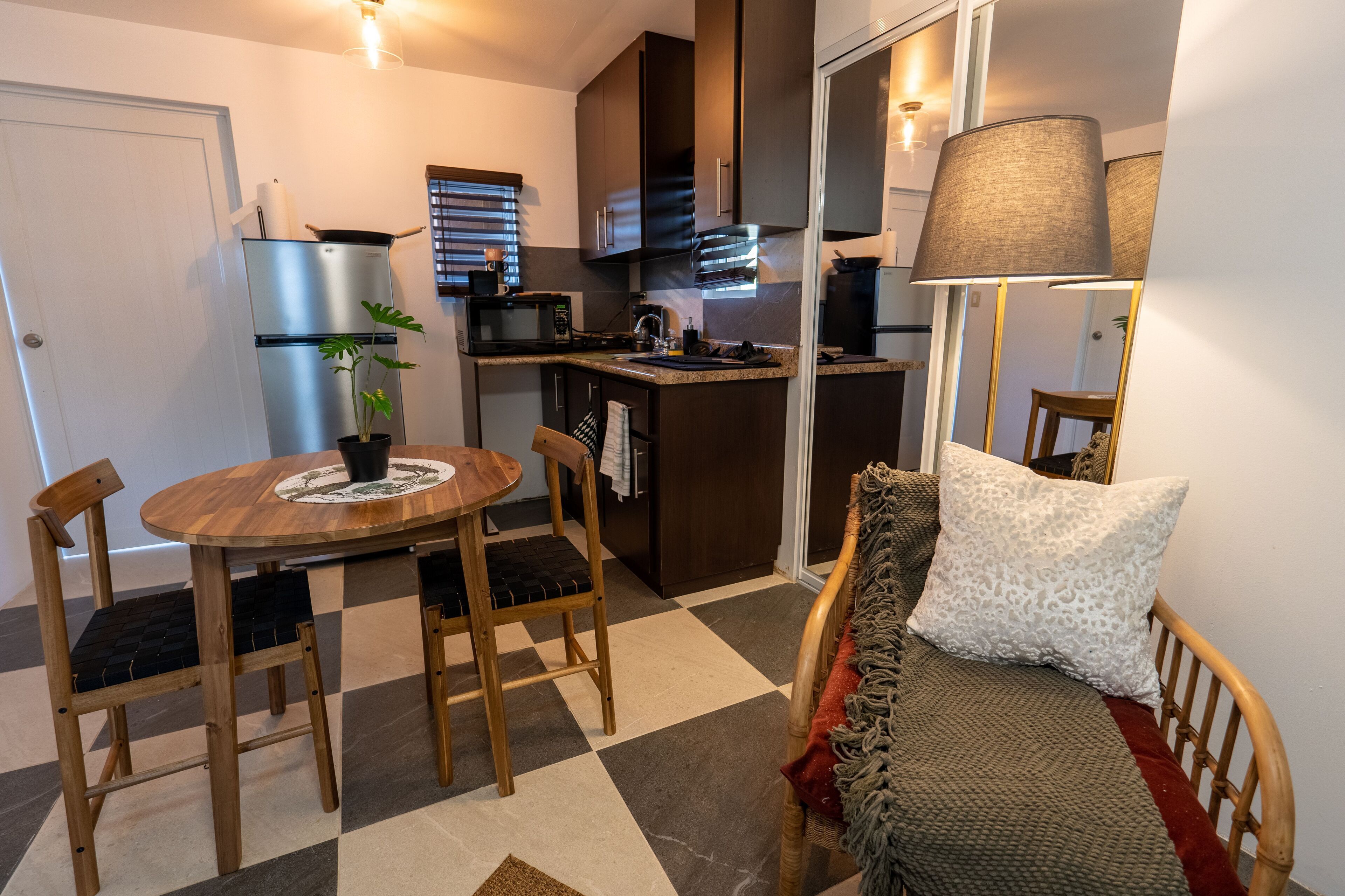 A compact kitchen with a refrigerator, microwave, and stove. There is a dining table with chairs and a small rug. A sofa with a throw blanket and pillows is visible.