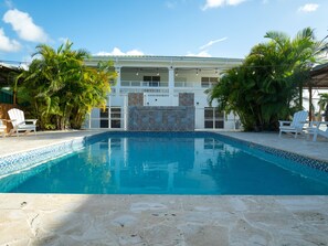 A swimming pool with a stone-tiled edge, surrounded by lounge chairs and palm trees, with a building featuring a balcony and stone wall in the background.