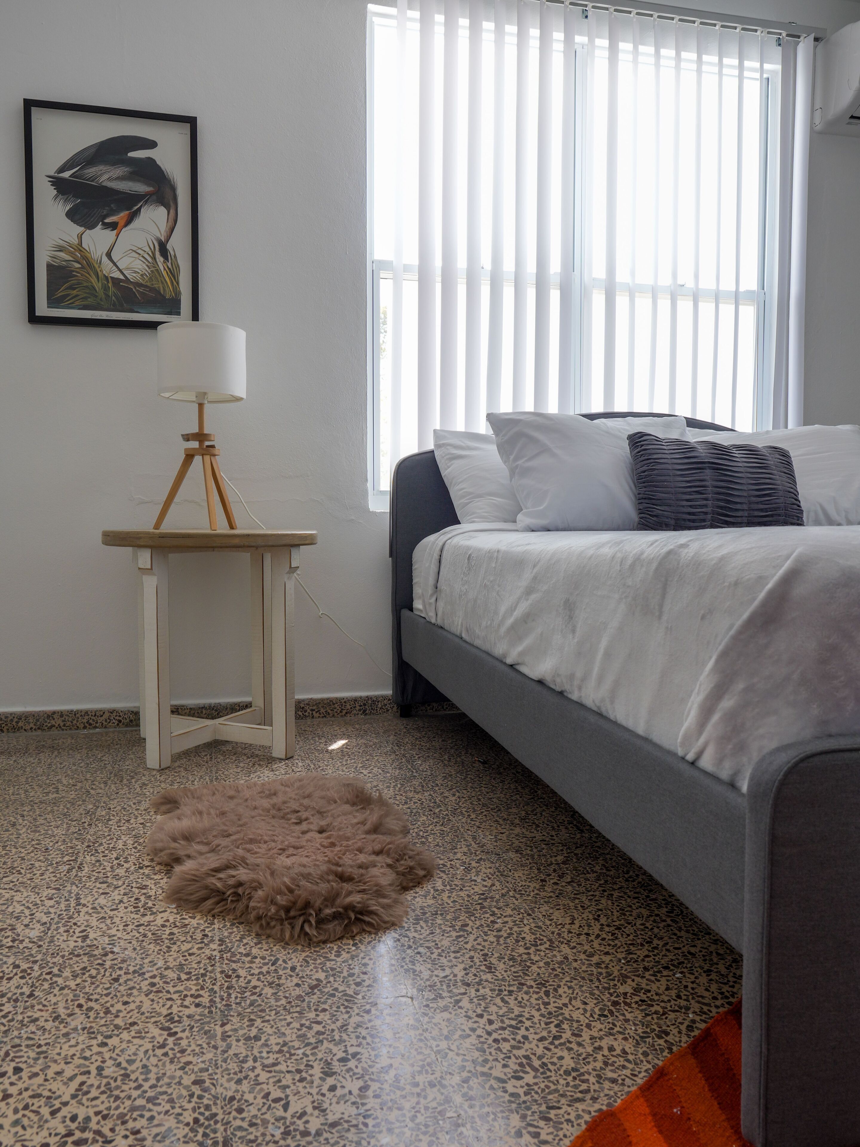 Ein Schlafzimmer mit einem Bett, einem kleinen Tisch mit Lampe, einem flauschigen Teppich, einem gerahmten Bild an der Wand und einem Fenster mit Jalousien.