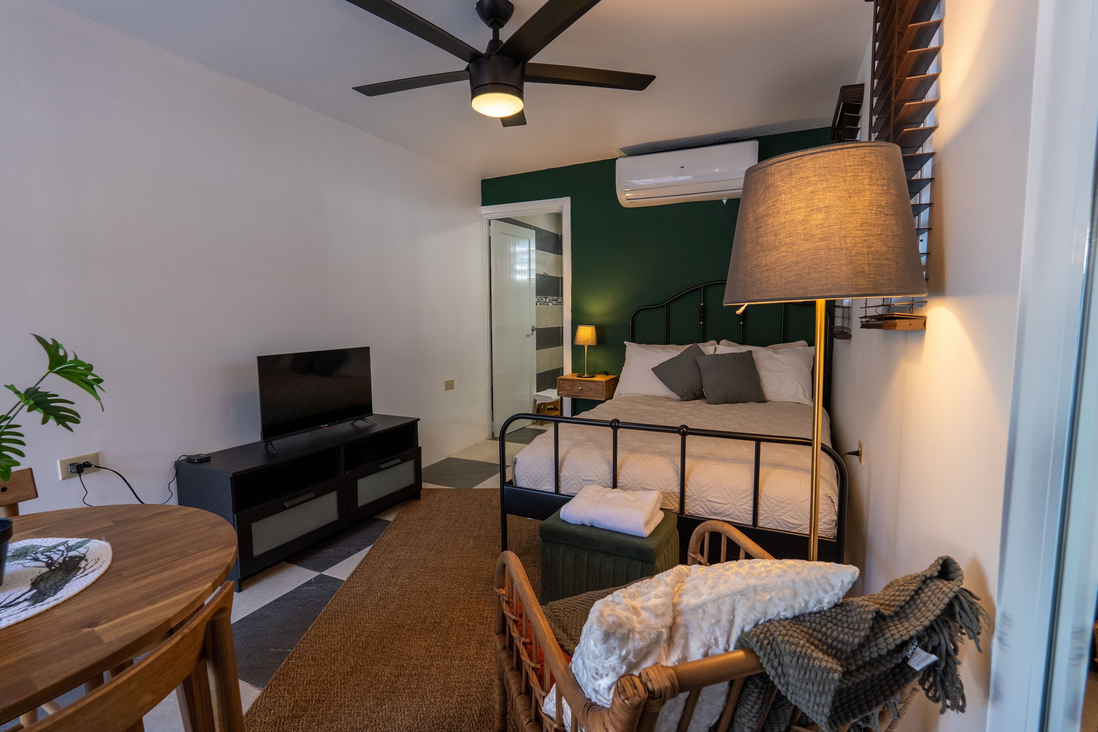Habitación de hotel pequeña con cama, televisión, ventilador de techo, mesa con sillas y un rincón comedor.