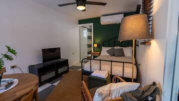 Habitación de hotel pequeña con cama, televisión, ventilador de techo, mesa con sillas y un rincón comedor.