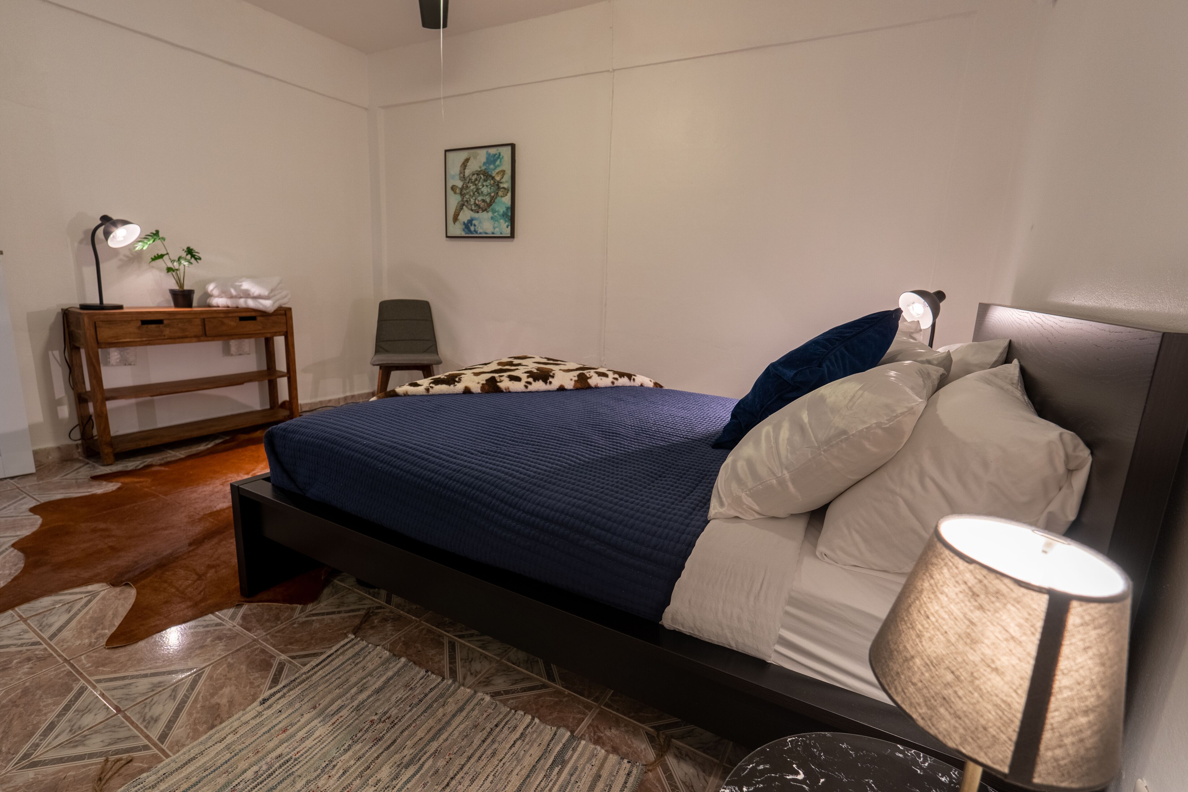 A bedroom with a bed, a nightstand, a lamp, a potted plant, and a painting on the wall.