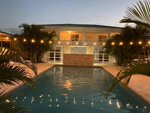 A pool area with a house in the background, surrounded by palm trees and string lights.