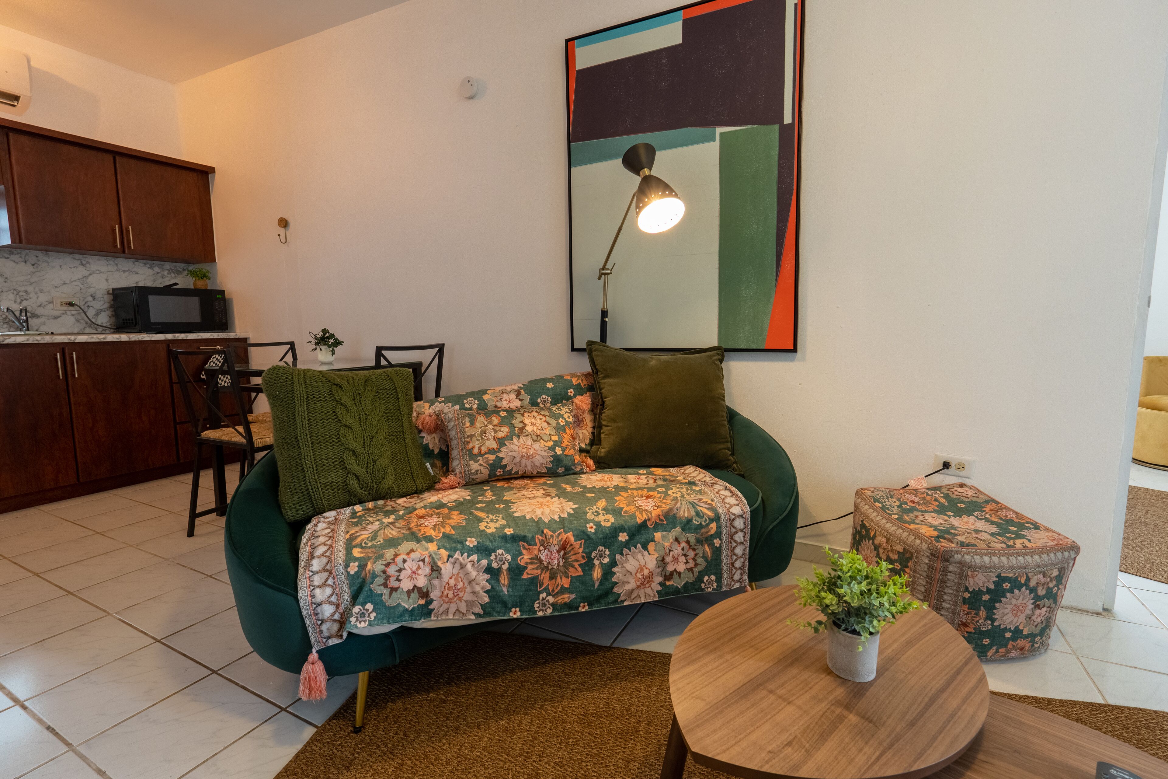 A living room with a sofa, a round coffee table, a small side table, a lamp, and a kitchen area with cabinets and a microwave.