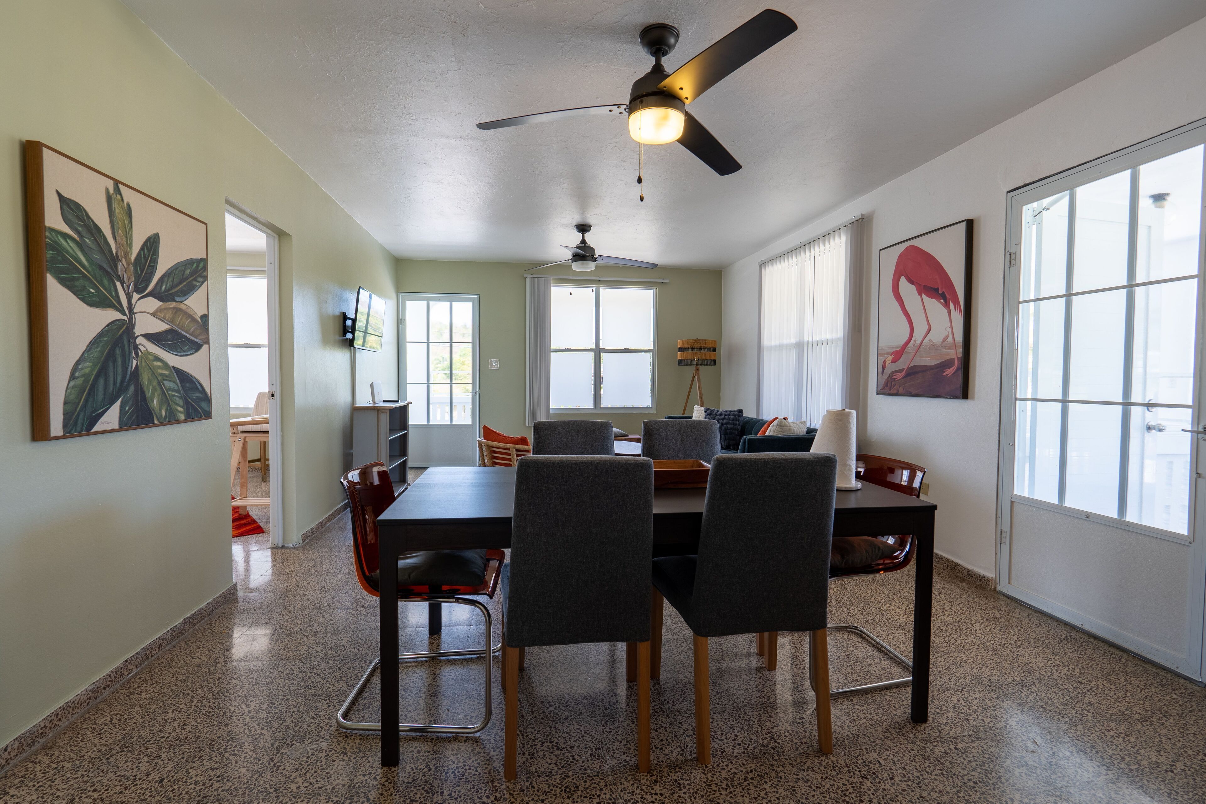 Uma sala de jantar com mesa, cadeiras, ventiladores de teto e janelas amplas.