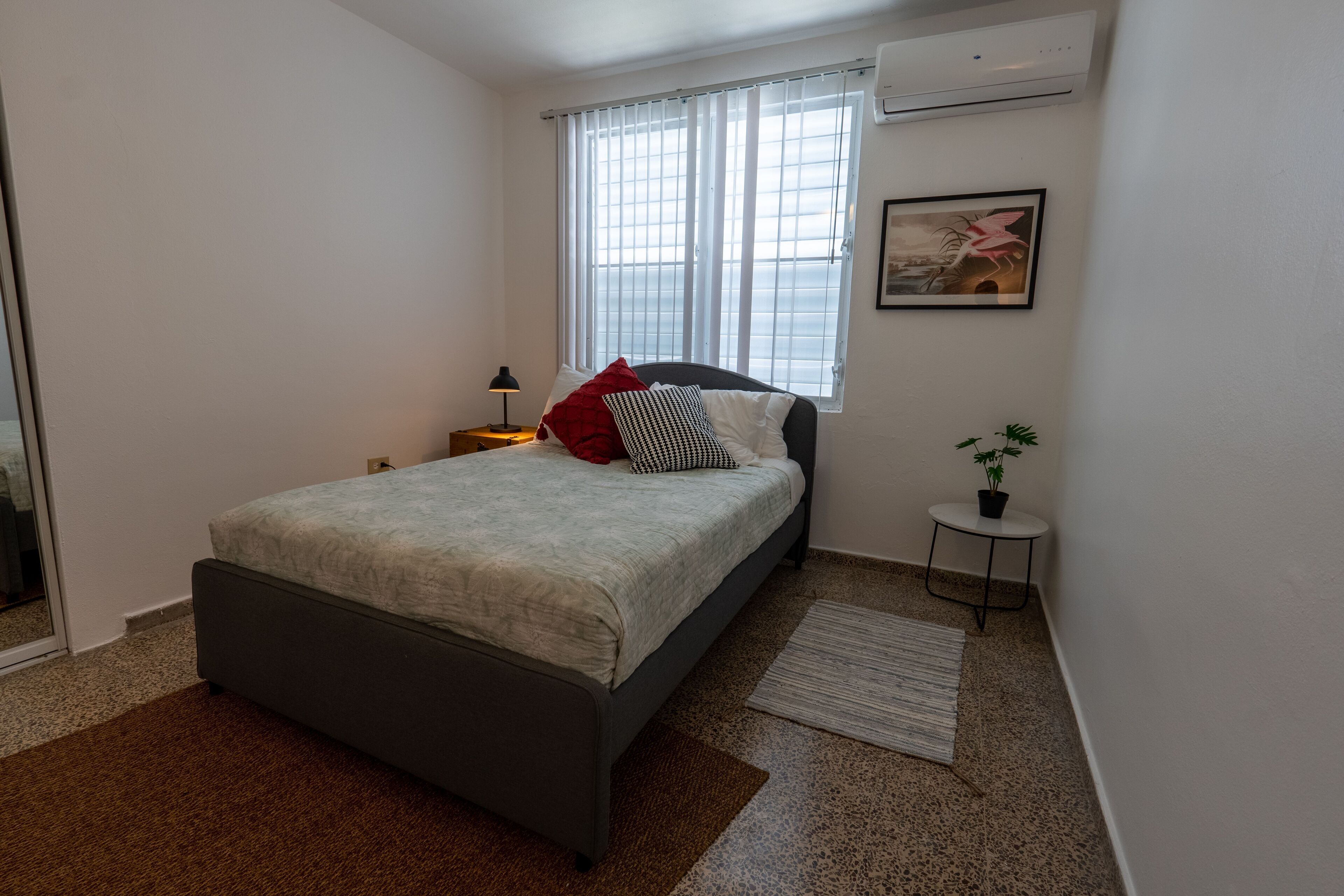 Ein Schlafzimmer mit einem Bett, einem Nachttisch, einem kleinen Tisch mit Pflanze, einem Fenster mit Jalousien und einer Wandklimaanlage.