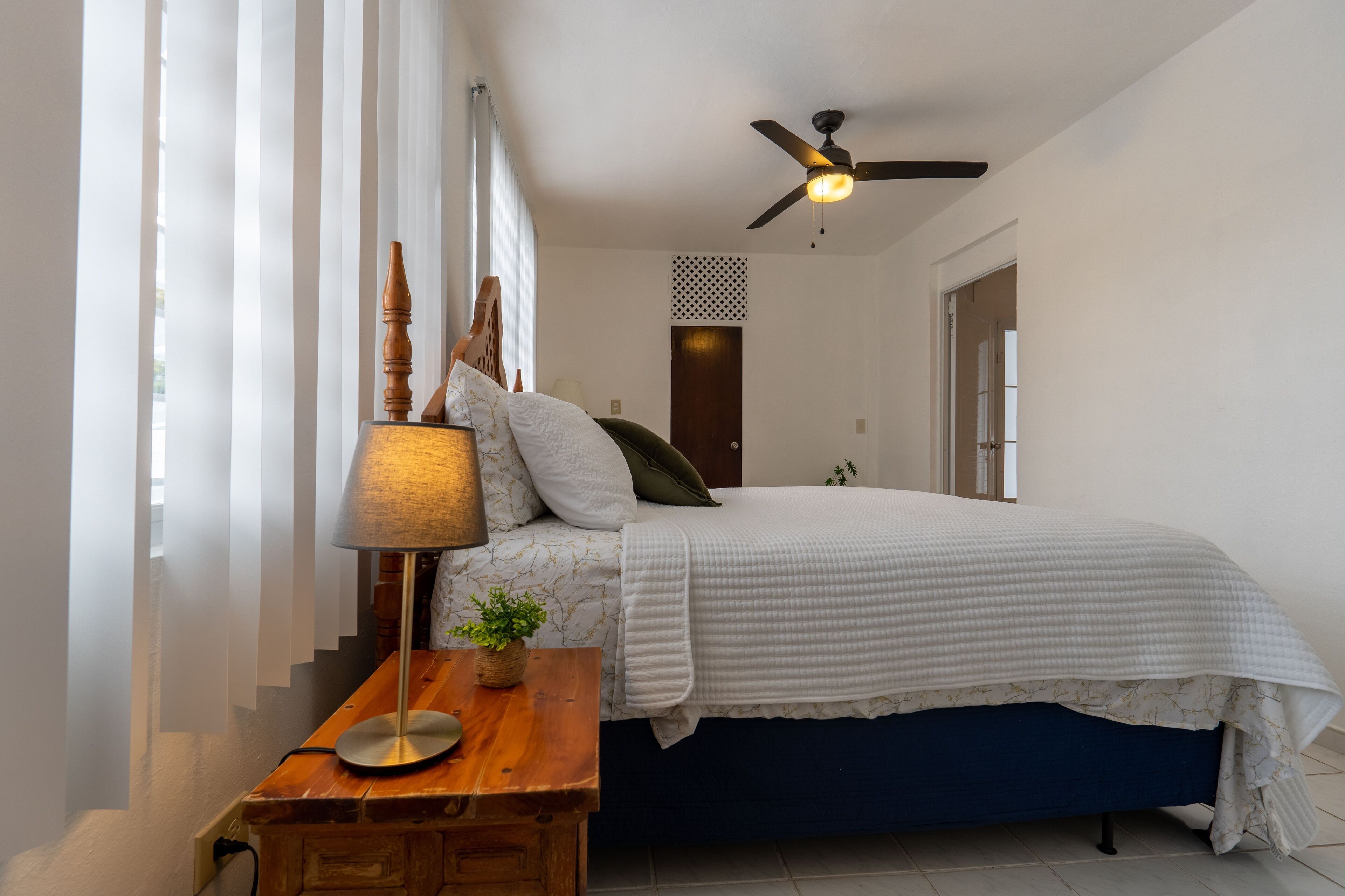 Una camera da letto con un letto, un comodino in legno, una lampada, una ventola a soffitto e una finestra con tende trasparenti.