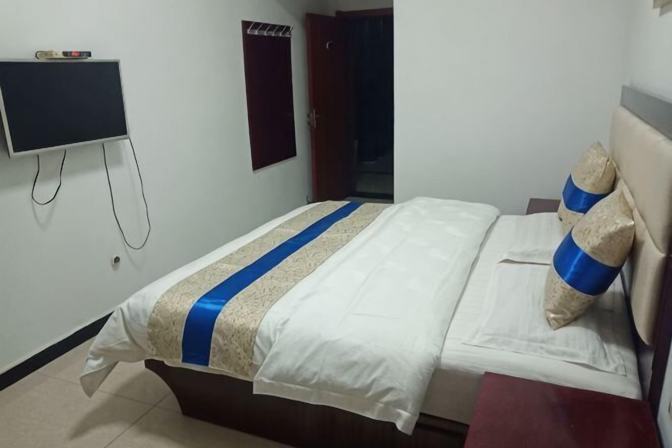 A neatly made bed with a white and blue bedspread, a television mounted on the wall, and a door in the background.