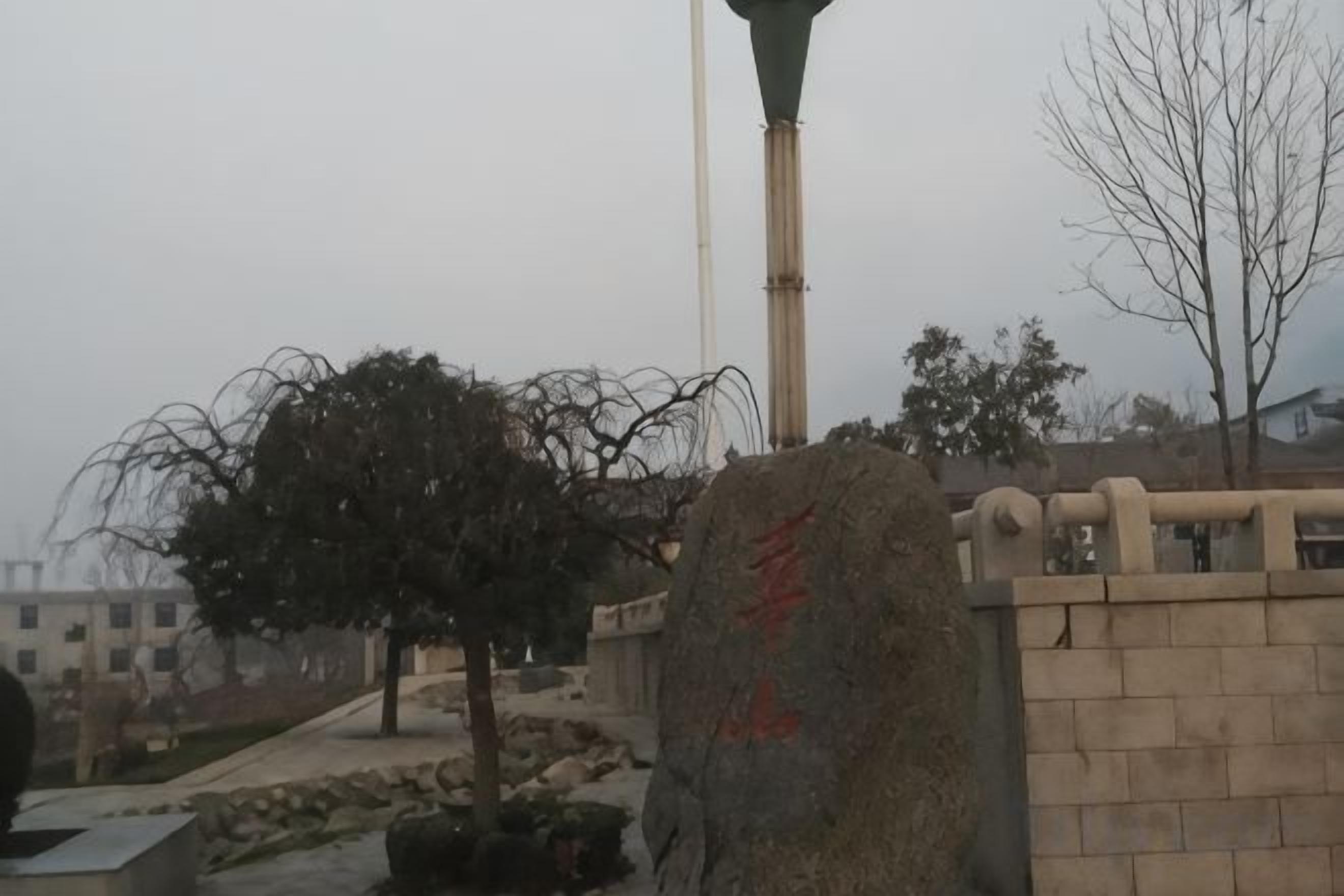 A stone wall with a large rock featuring red text, a lamp post, and a leafless tree.