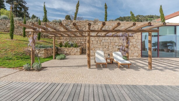 A wooden pergola with two white lounge chairs and a small table, set against a stone wall with a view of trees and a modern building. - Quinta do Pinhô - Tarouca (Tarouca)