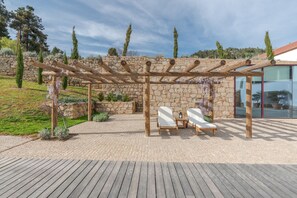 A wooden pergola with two white lounge chairs and a small table, set against a stone wall with a view of trees and a modern building. - Quinta do Pinhô - Tarouca (Tarouca)