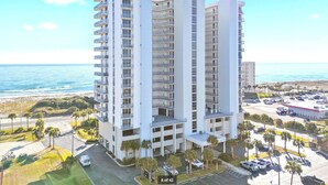 A high-rise hotel with multiple floors, a central tower, and a parking area with cars and palm trees. - 10th Floor Sweeping Views of the Gulf. Steps from Beach Access. (Pensacola Beach)
