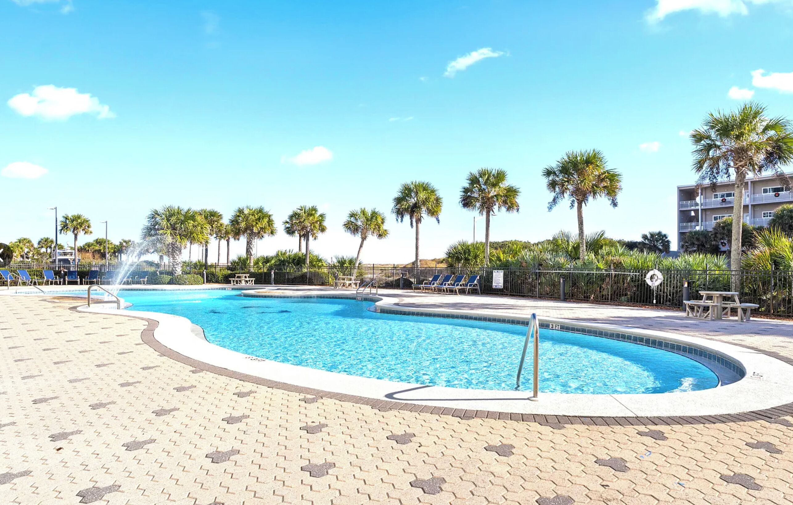 A swimming pool with lounge chairs and palm trees, surrounded by a paved area and a fence.