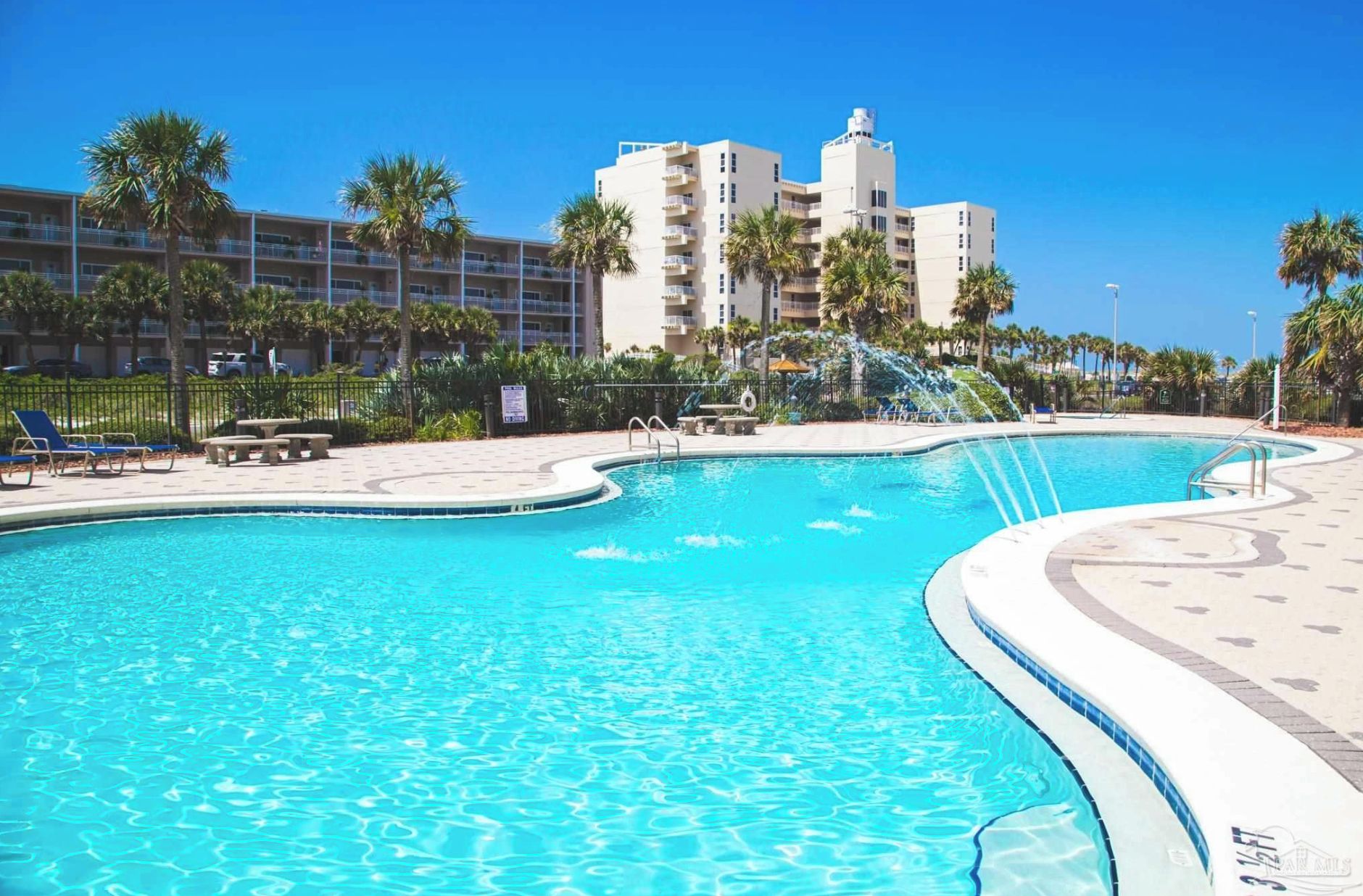 A swimming pool with a curved design, surrounded by palm trees and a building with multiple balconies.