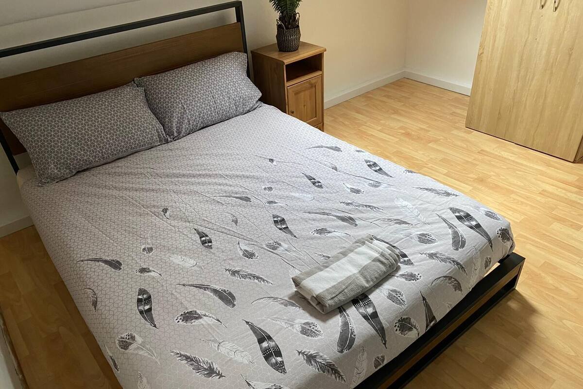 Ein Bett mit einem geometrisch gemusterten Überwurf, einem Holzkopfende und einem Nachttisch mit einer Topfpflanze.