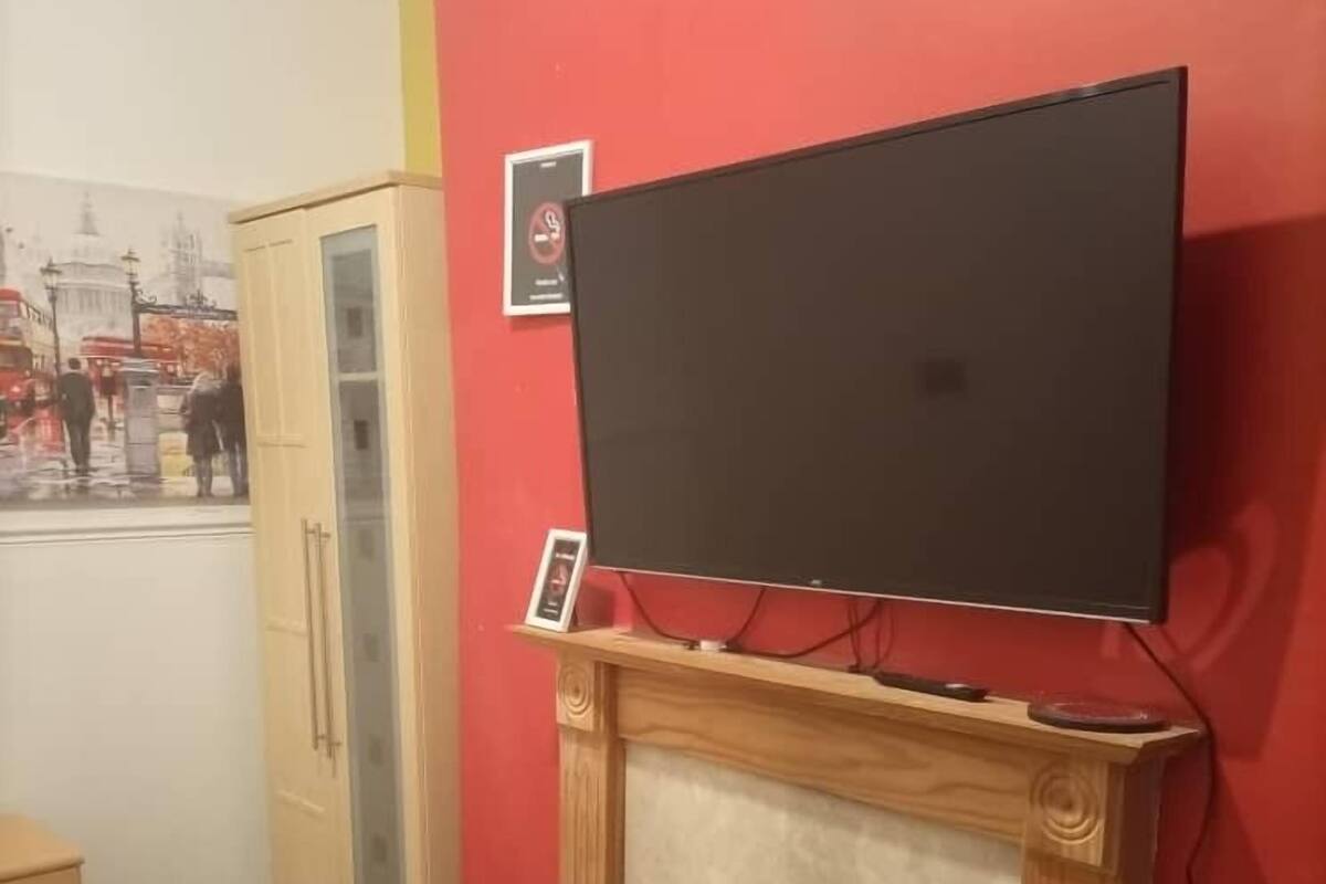 Ein Zimmer mit roter Wand, einem flachbildfernseher über einem Kamin, einem Holzschrank und einem gemusterten Hocker.