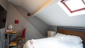 Une chambre avec un plafond en pente, un lit, un bureau avec une chaise, une table de chevet et un ventilateur fixé au mur.
