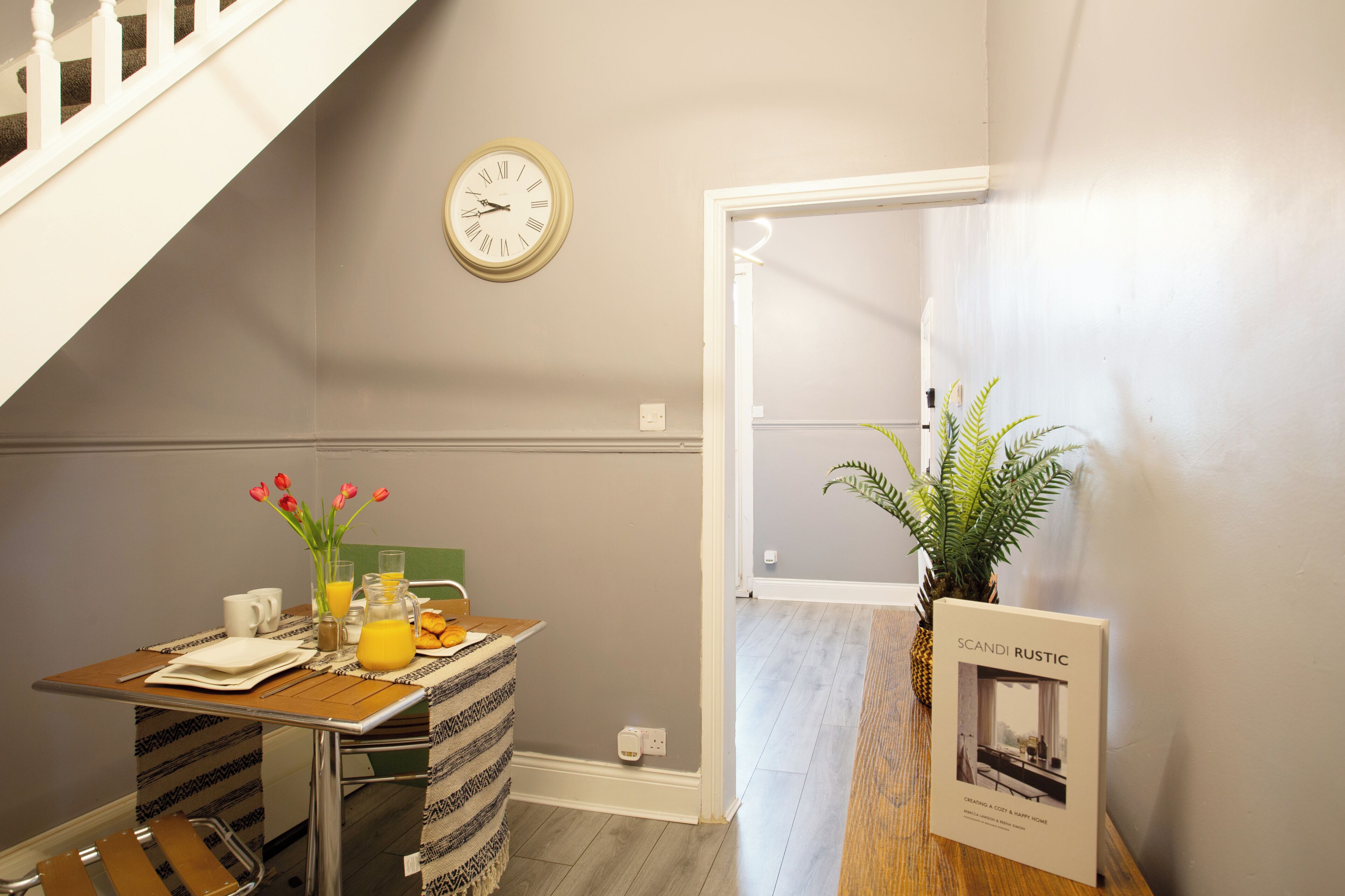 Ein Zimmer mit Esstisch, gedeckt für das Frühstück, einer Uhr an der Wand und einer Pflanze neben der Tür.