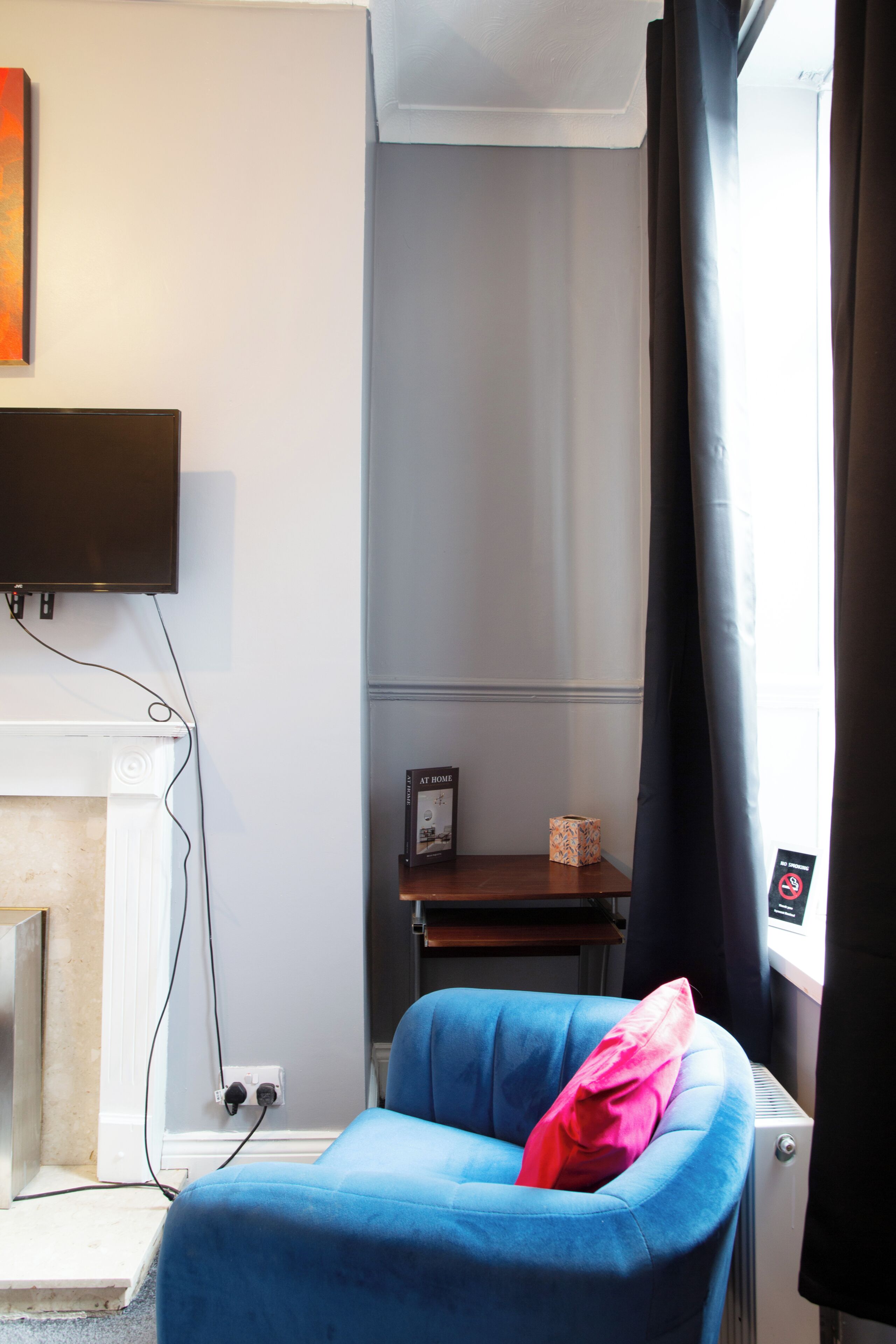 A room with a blue chair, a flat-screen TV mounted on the wall, a fireplace, and a window with curtains.
