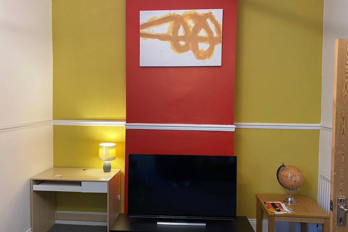 Ein Zimmer mit Fernseher auf schwarzem Standfuß, einem kleinen Tisch mit Lampe und einem Globus auf einem Holznachttisch.