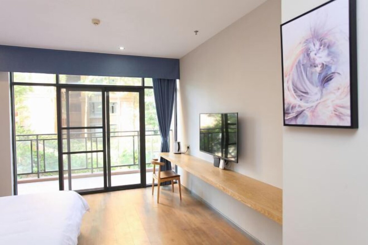 A modern hotel room with a large sliding glass door to a balcony, a bed, a wooden chair, a flat-screen TV mounted on the wall, and a framed artwork.