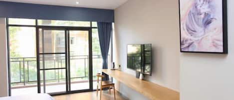 Une chambre d’hôtel moderne dotée d’une grande porte-fenêtre coulissante donnant sur un balcon, d’un lit, d’une chaise en bois, d’un téléviseur à écran plat fixé au mur et d’une œuvre d’art encadrée.