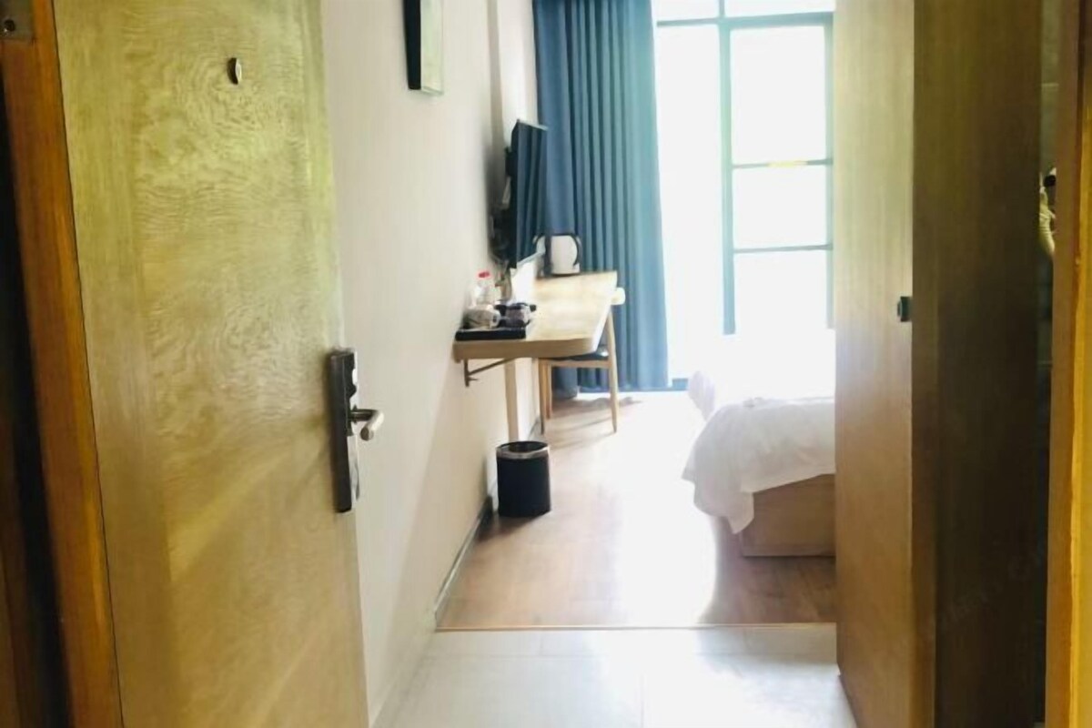 A hotel room with a wooden door, a small desk with a computer and a trash bin, a bed with white bedding, and a window with blue curtains.