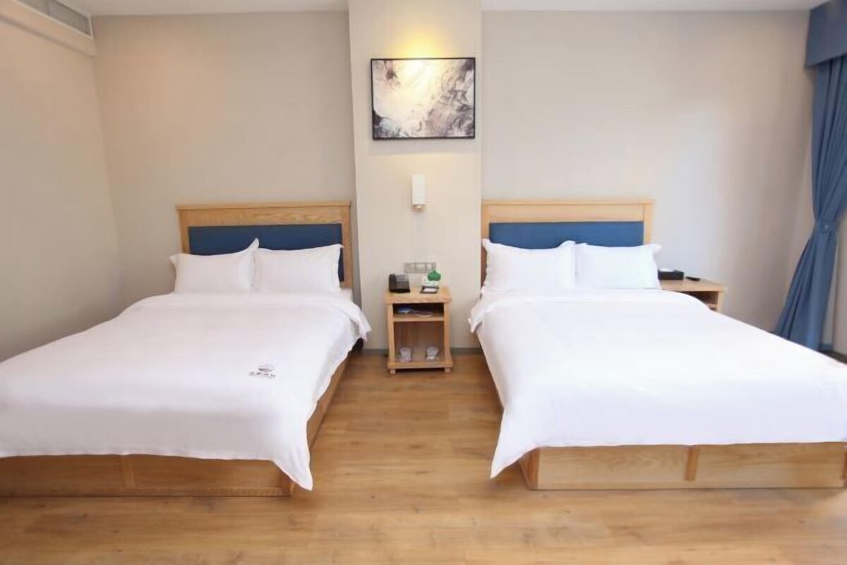 A hotel room with two single beds, wooden headboards, white bedding, a small bedside table with a lamp, and a wall-mounted artwork.