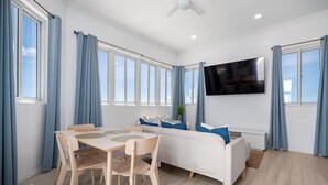 Ein modernes Zimmer mit Esstisch, Stühlen, einer Couch, einem Fernseher und großen Fenstern mit Vorhängen.