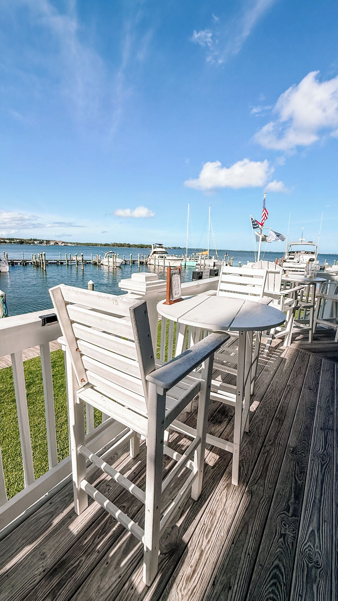 A wooden deck with white furniture overlooking a marina with boats and a clear blue sky.