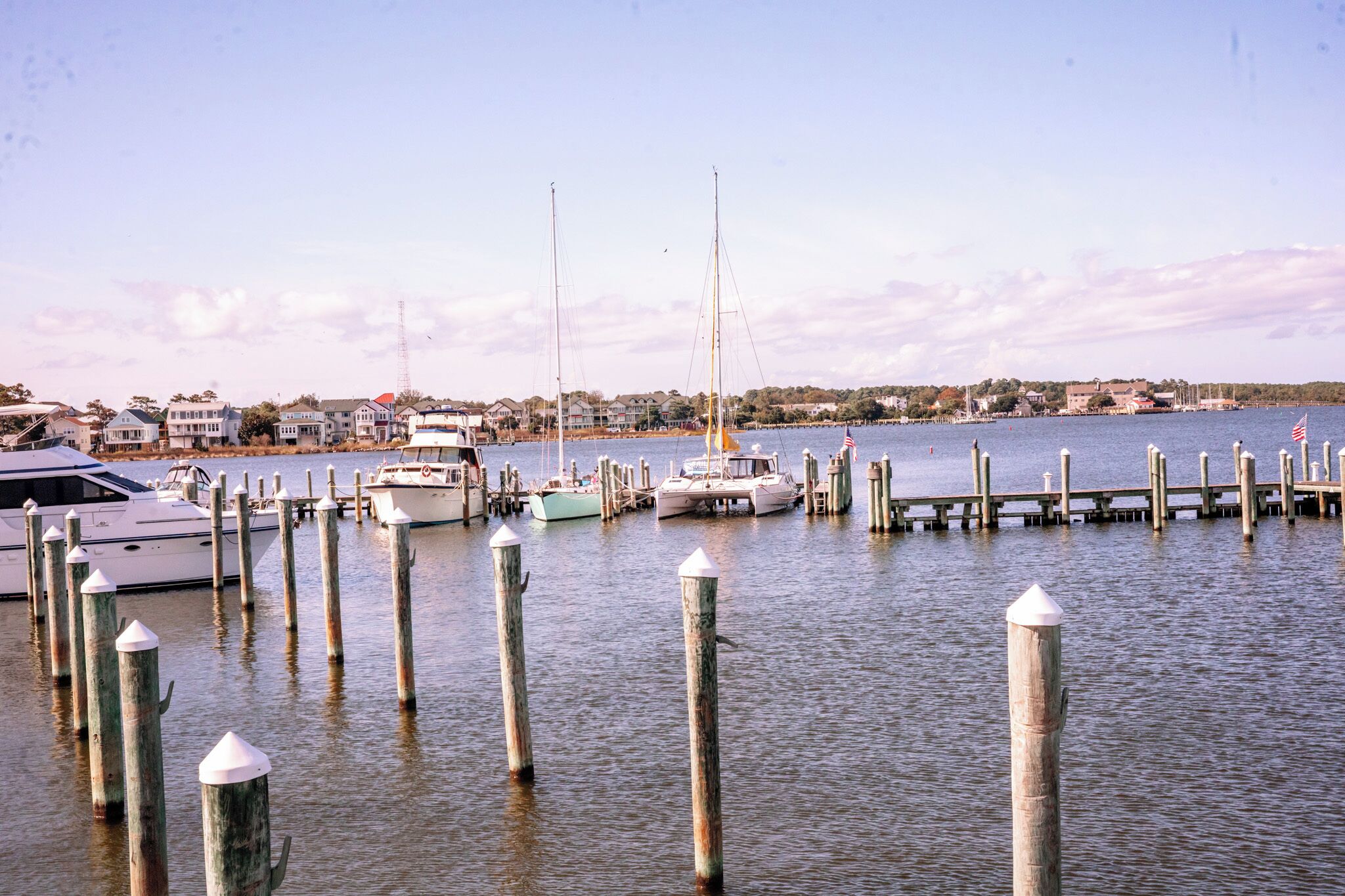 A marina with boats docked at wooden piers, a clear sky, and buildings in the background.