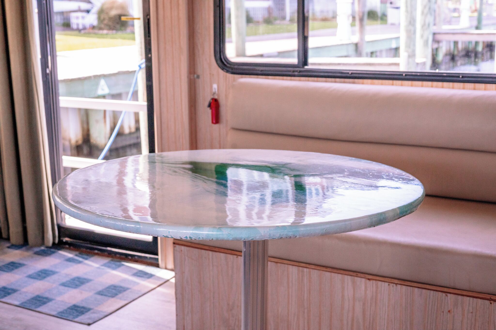 Una mesa redonda de vidrio con base metálica, ubicada frente a un banco con cojín color beige.