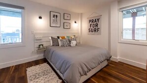 A bedroom with a bed, a bedside table, a rug, and a wall with framed pictures and a 'PRAY FOR SURF' sign. - The Recovery Room-0.5 Mi to Beach & 0.3 to Sound (Kill Devil Hills)