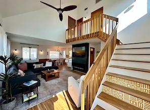 A spacious living area with a large TV, a wooden staircase, and a ceiling fan.