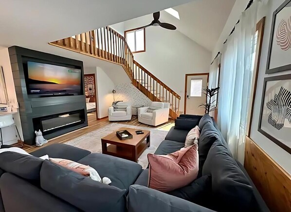 A modern living room with a large flat-screen TV, a staircase, and a cozy seating area. - Knot On Call: OceanViews/Pool/MultiUsePath (Kill Devil Hills)