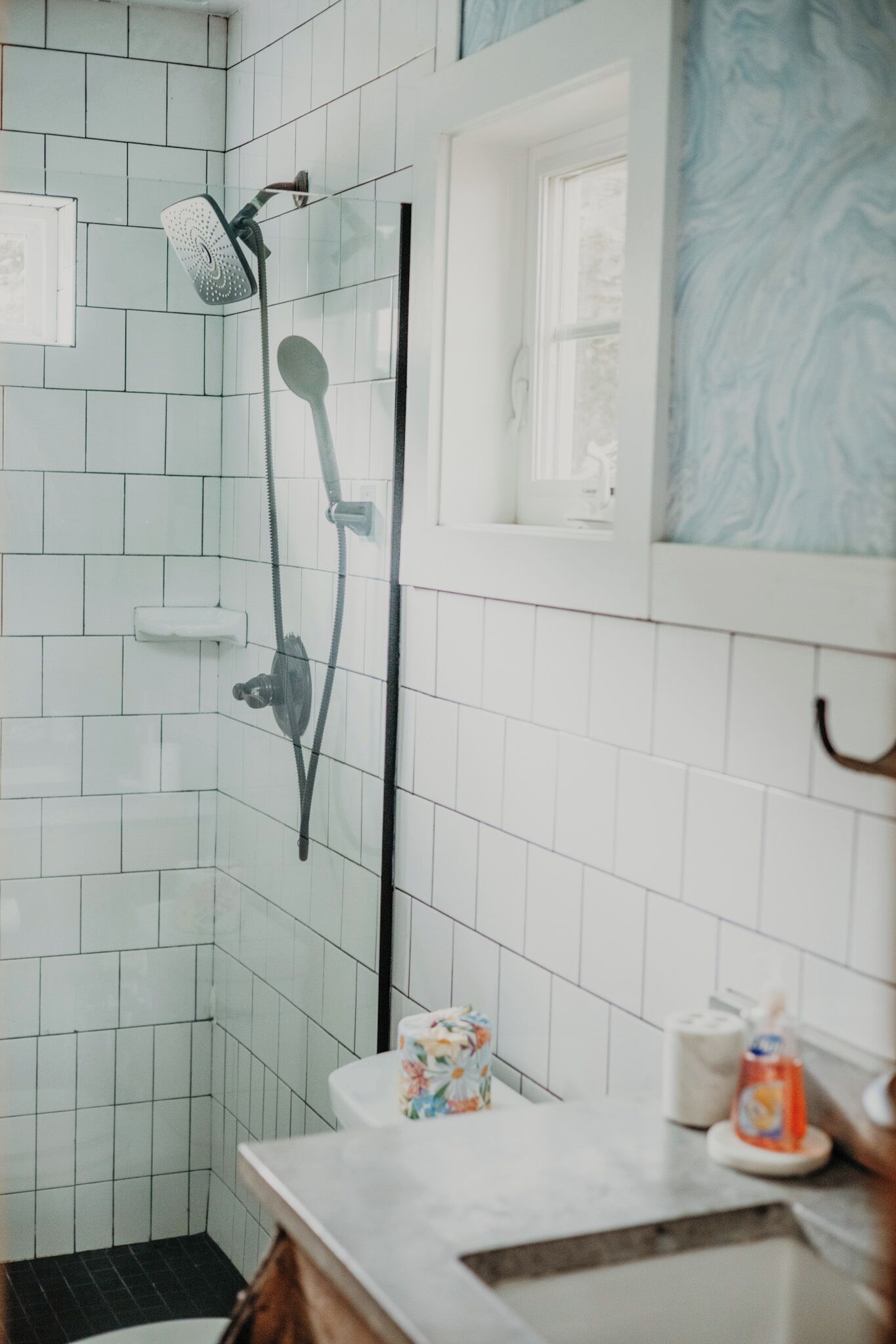 Un baño con paredes de azulejos blancos, una ducha con cabezal de ducha de mano, un inodoro con dispensador de toallas de papel con motivos florales y una ventana con diseño de mármol azul.