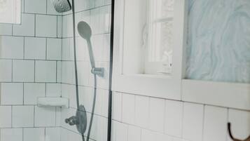 Un baño con paredes de azulejos blancos, una ducha con cabezal de ducha de mano, un inodoro con dispensador de toallas de papel con motivos florales y una ventana con diseño de mármol azul.