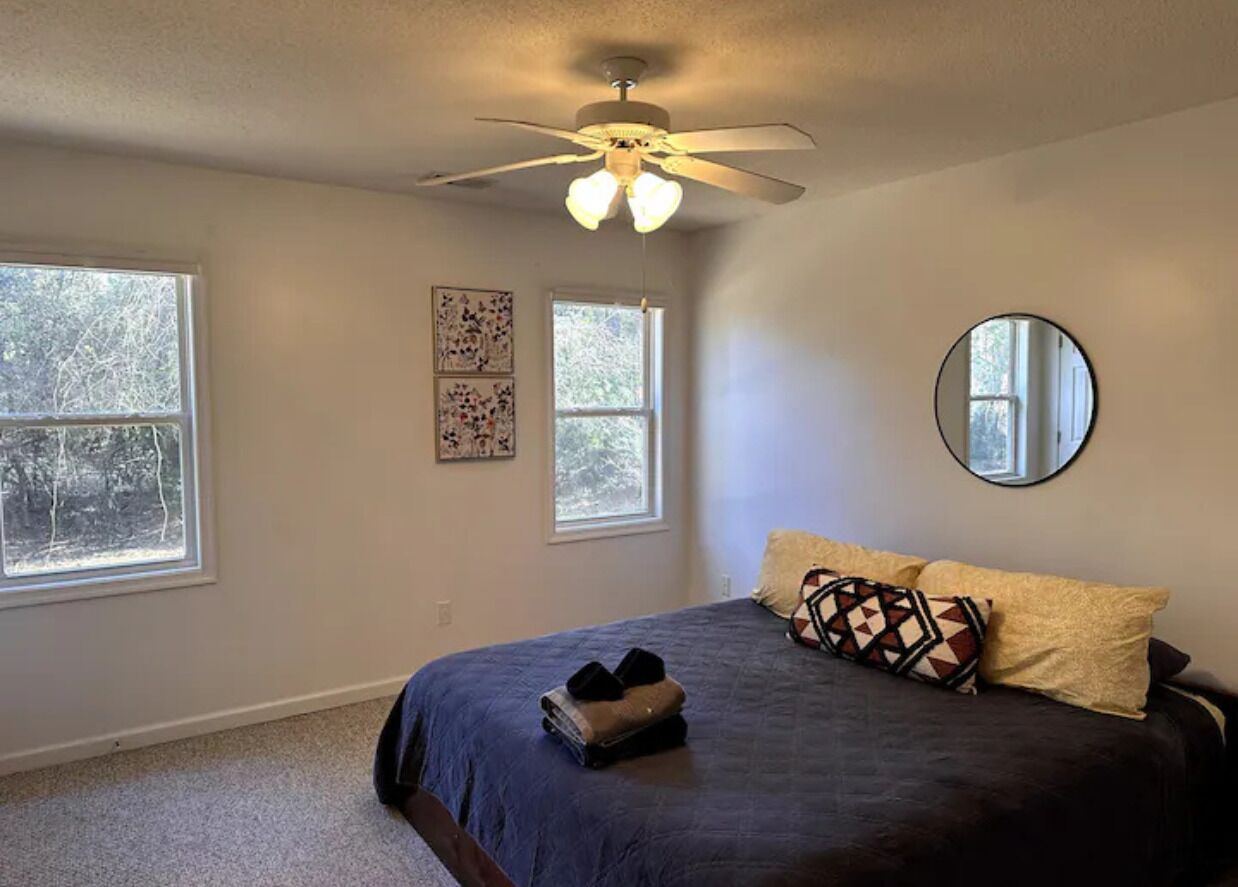 Un dormitorio con cama, ventilador de techo, dos ventanas y un espejo.