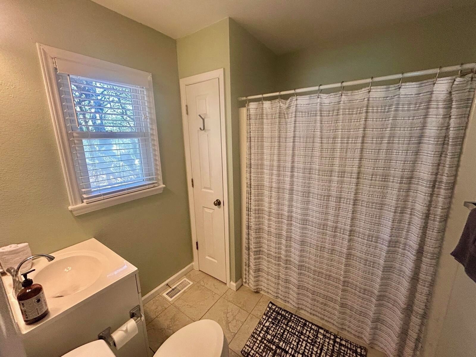 Une salle de bain comprenant des toilettes, un lavabo, une fenêtre avec stores, un rideau de douche et un tapis.