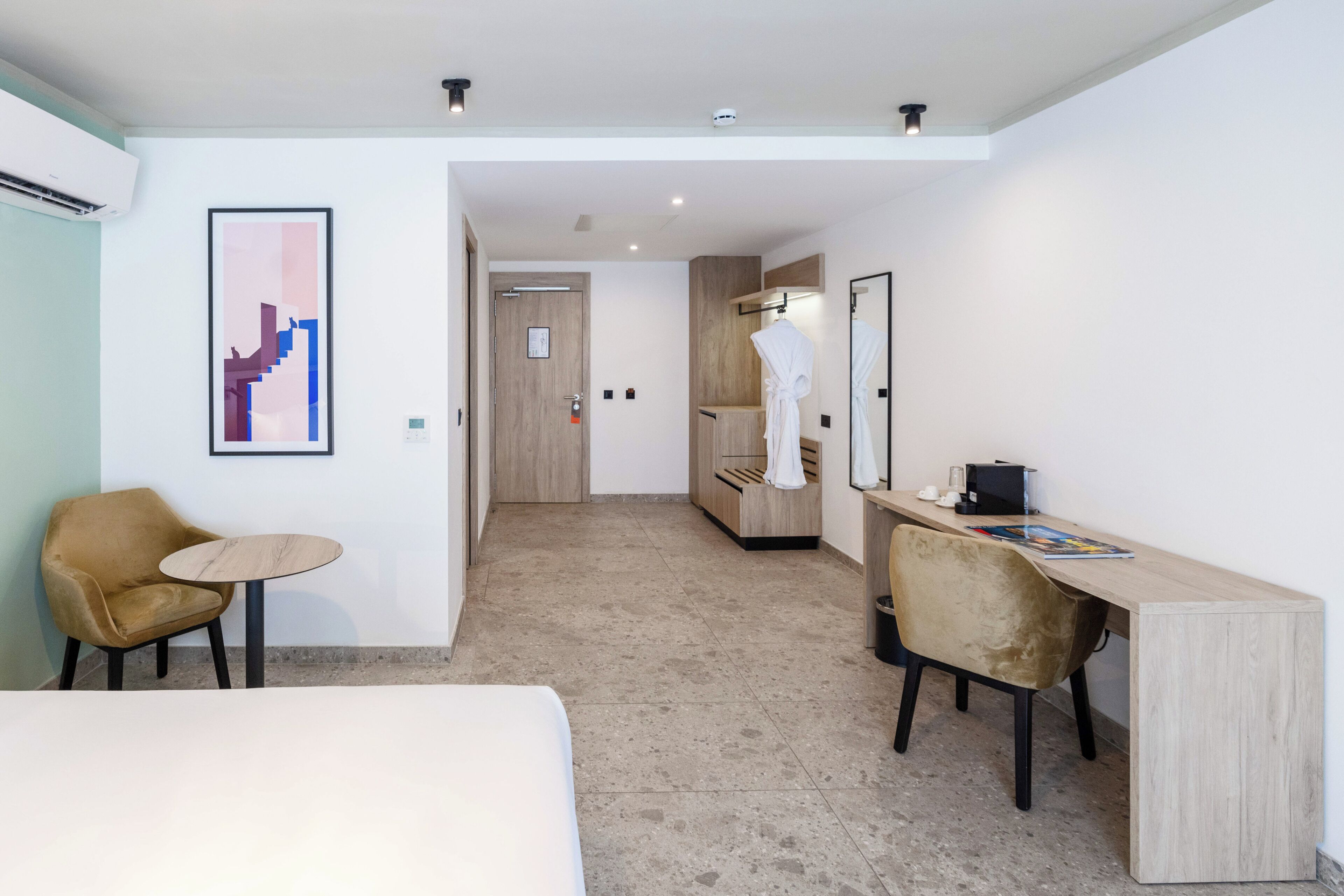 A modern hotel room with a bed, a desk, a chair, and a small table. There is a bathroom with a mirror and a robe hanging on the door.